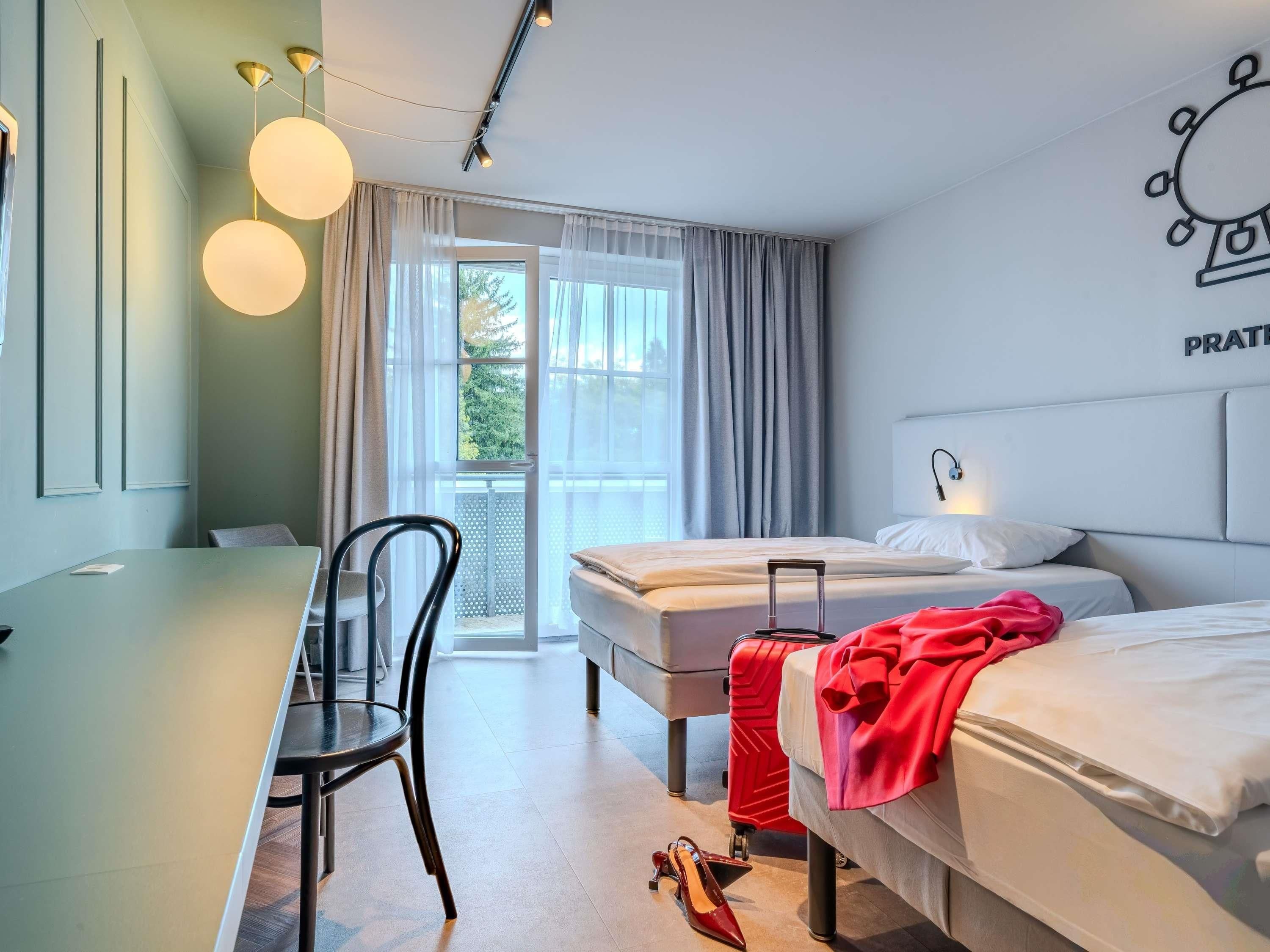 Habitación de hotel con dos camas, un escritorio, una silla, una maleta y una ventana con cortinas.