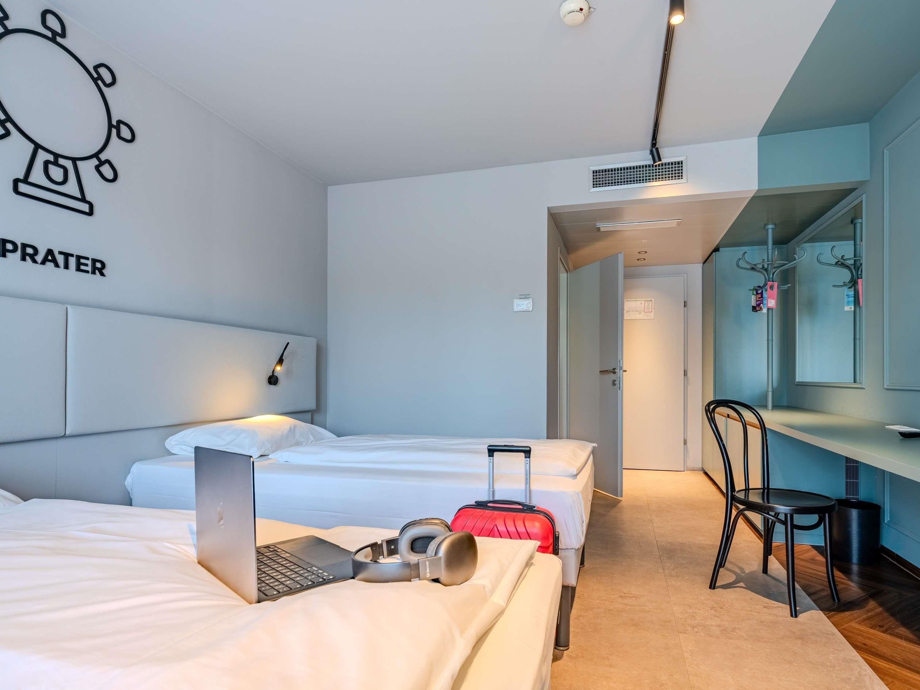 A hotel room with two beds, a desk, a chair, a laptop, and a red suitcase.
