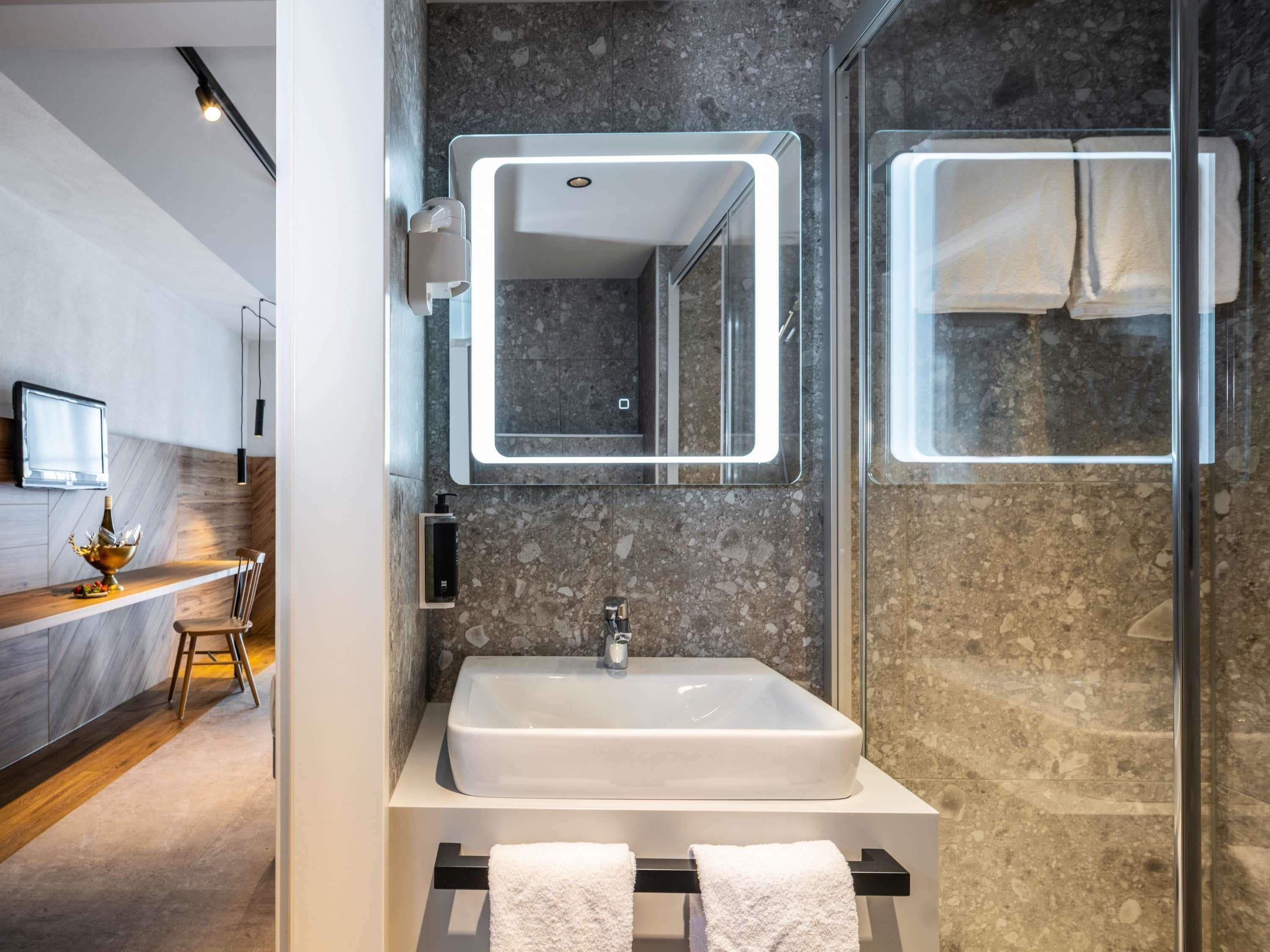 A modern bathroom with a large sink, a mirror with integrated lighting, a glass shower enclosure, and a wooden vanity with a bottle and glasses.
