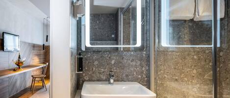 Un baño moderno con un lavamanos grande, un espejo con iluminación integrada, una cabina de ducha de cristal y un mueble de tocador de madera con una botella y vasos.