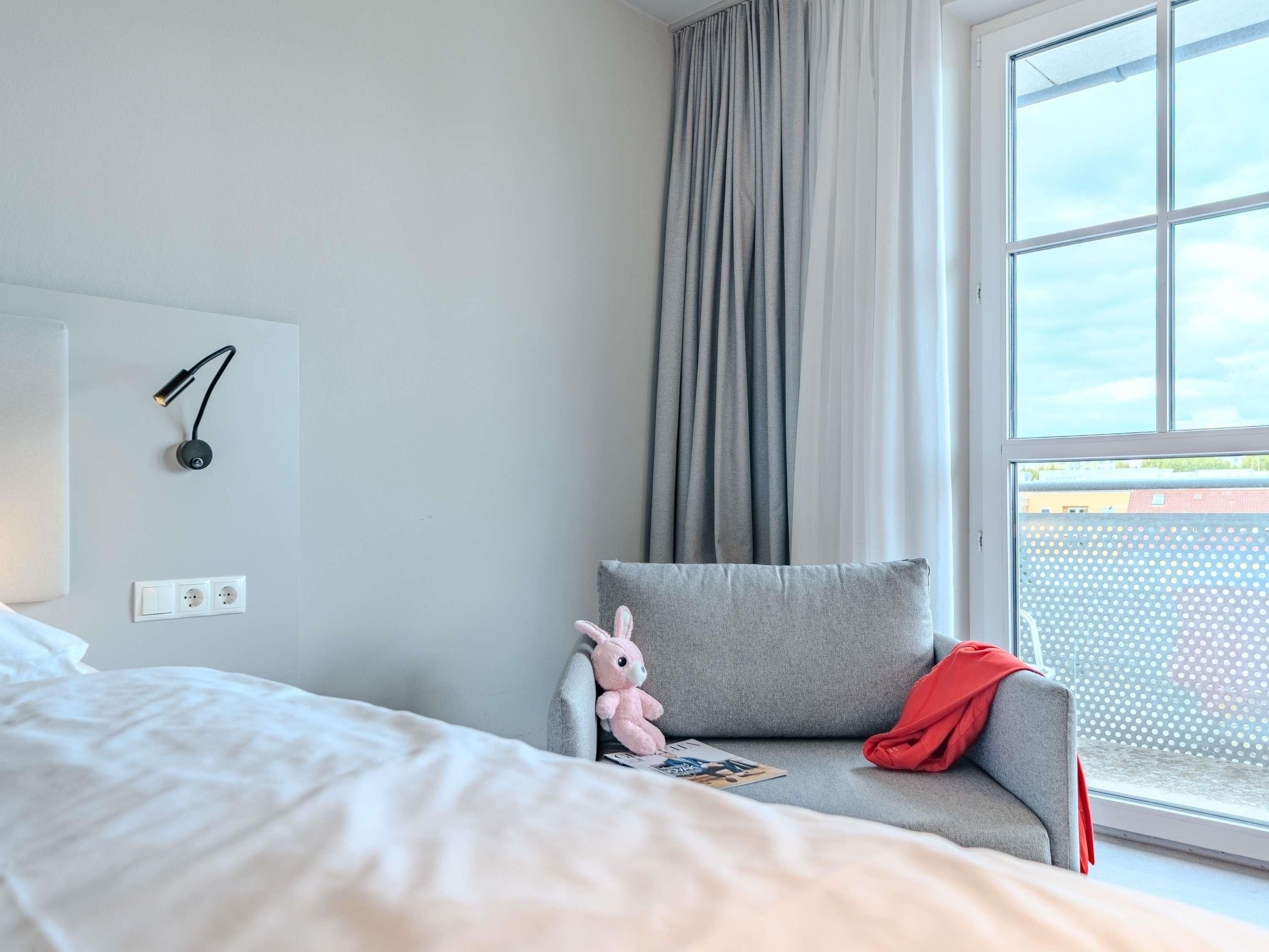 A hotel room with a bed, a gray armchair with a red throw, a stuffed toy, a wall-mounted lamp, and a window with a view.