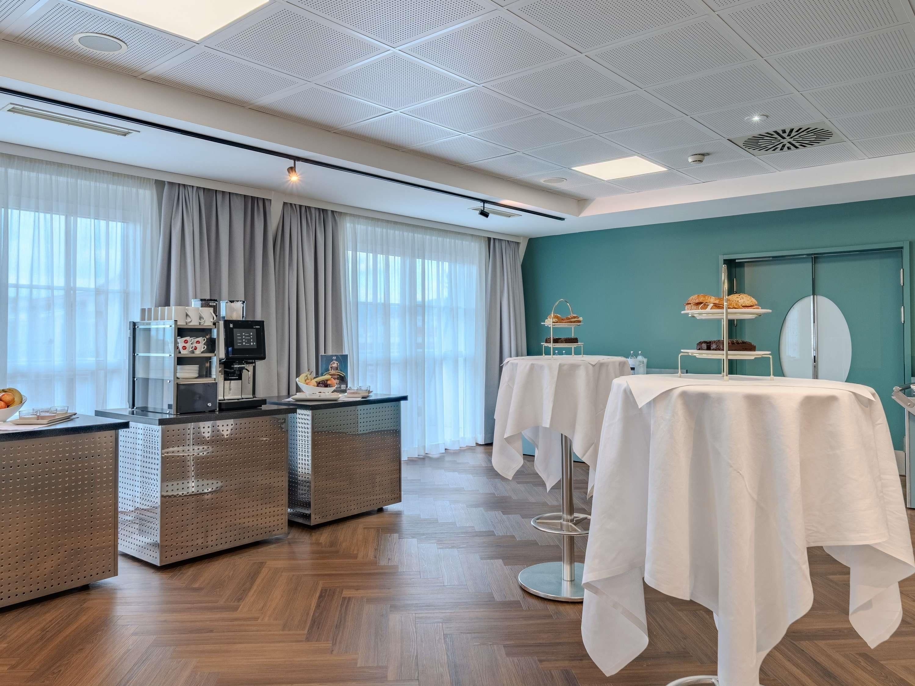 A hotel room with a kitchenette, a dining area with tables, a teal accent wall, and a large window with curtains.