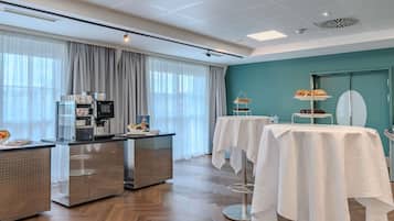 A hotel room with a kitchenette, a dining area with tables, a teal accent wall, and a large window with curtains.