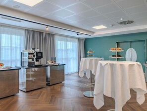 A hotel room with a kitchenette, a dining area with tables, a teal accent wall, and a large window with curtains. - Ibis Styles Graz Messe (Graz)