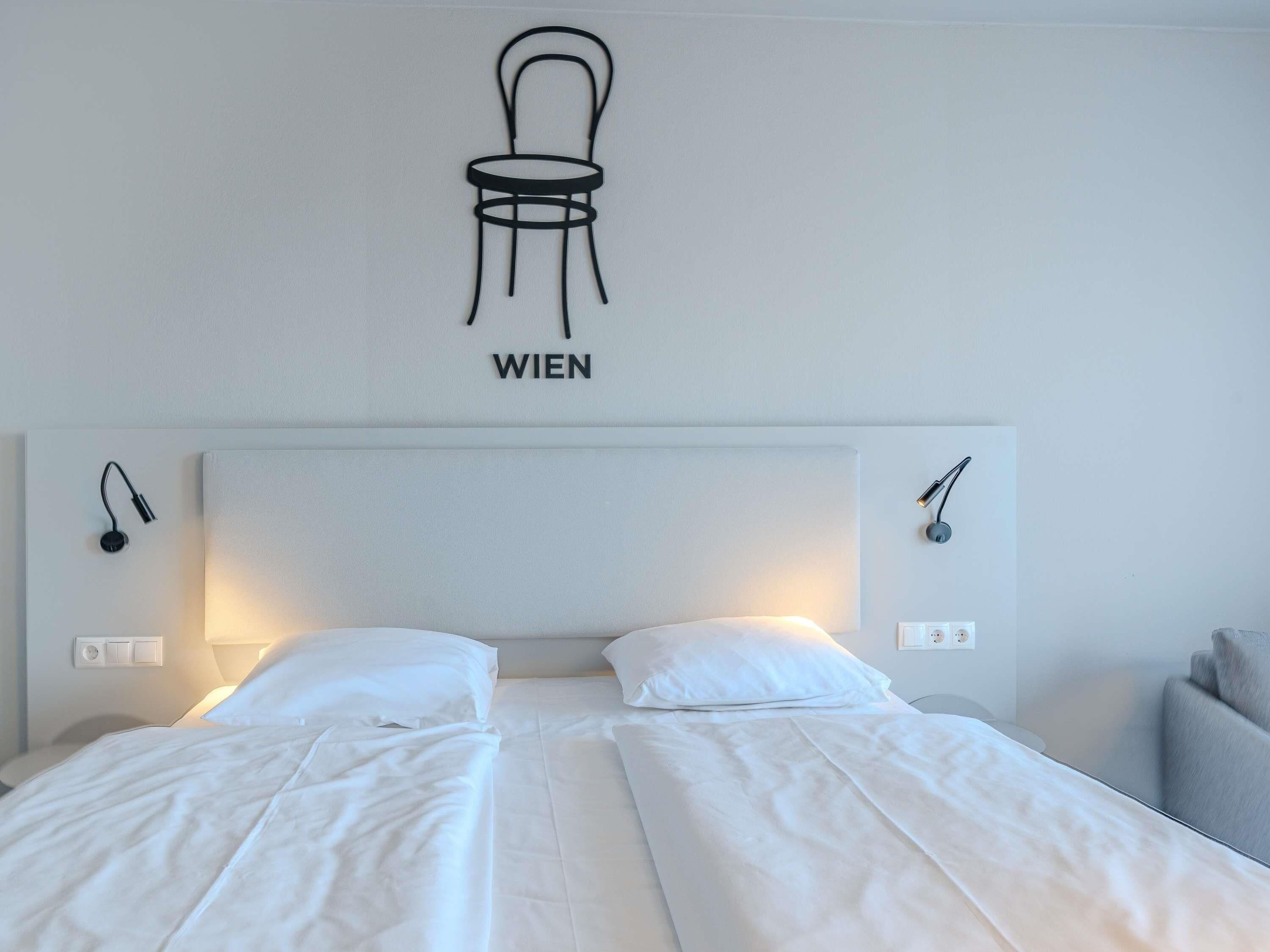 Ein Hotelzimmer mit einem Bett, zwei Nachttischen, einer Wandlampe und einer Wanddekoration, die einen Stuhl mit dem Wort 'WIEN' zeigt.
