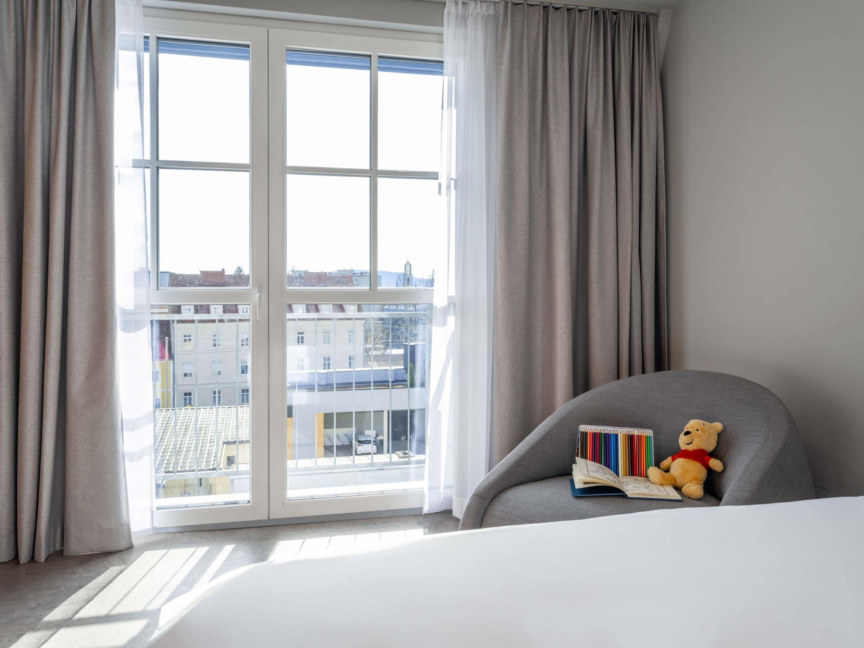 A hotel room with a large window, a grey armchair, and a teddy bear.