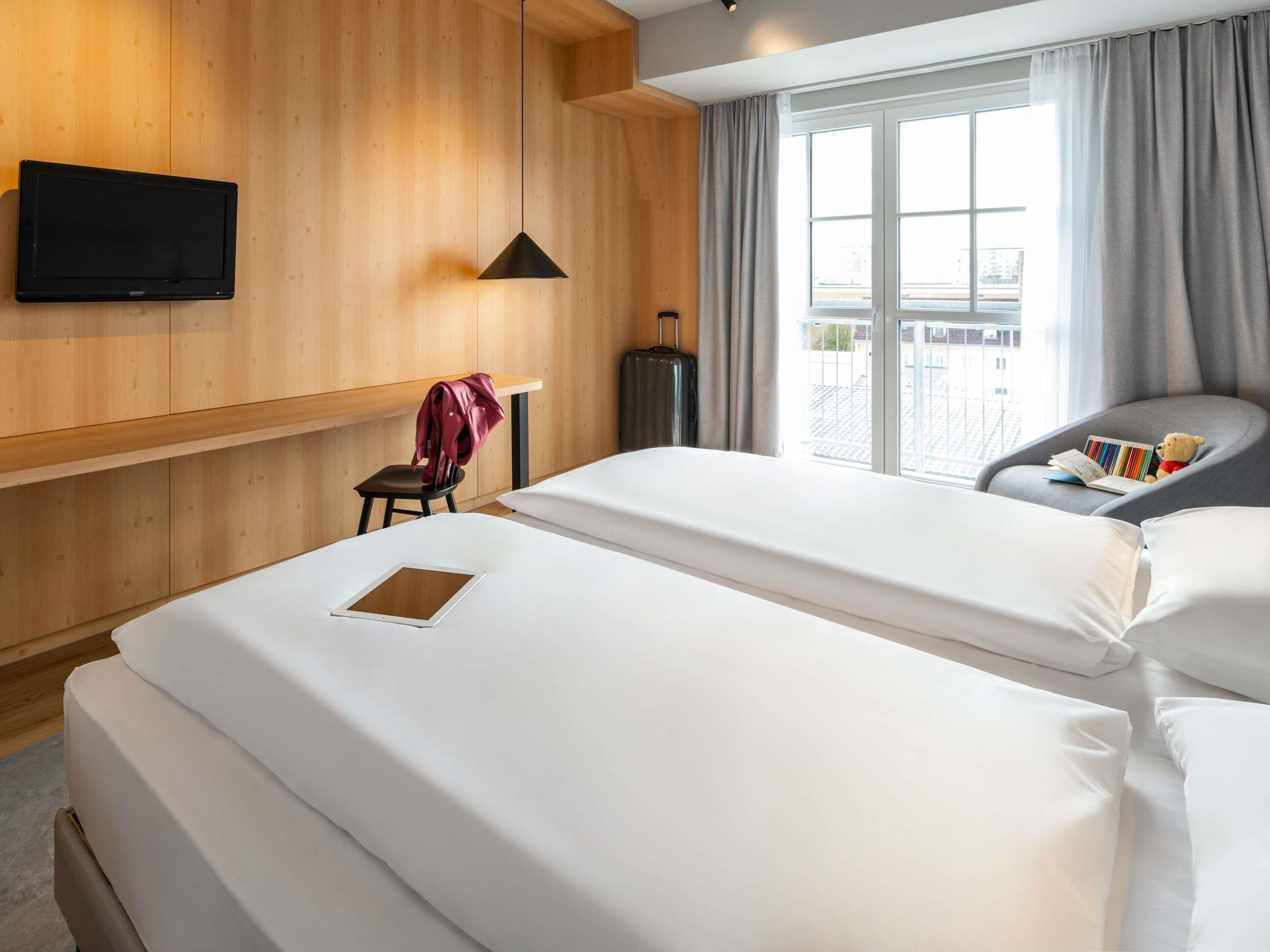 A hotel room with two beds, a TV mounted on the wall, a chair with a teddy bear, and a window with curtains.