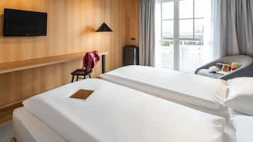 A hotel room with two beds, a TV mounted on the wall, a chair with a teddy bear, and a window with curtains.