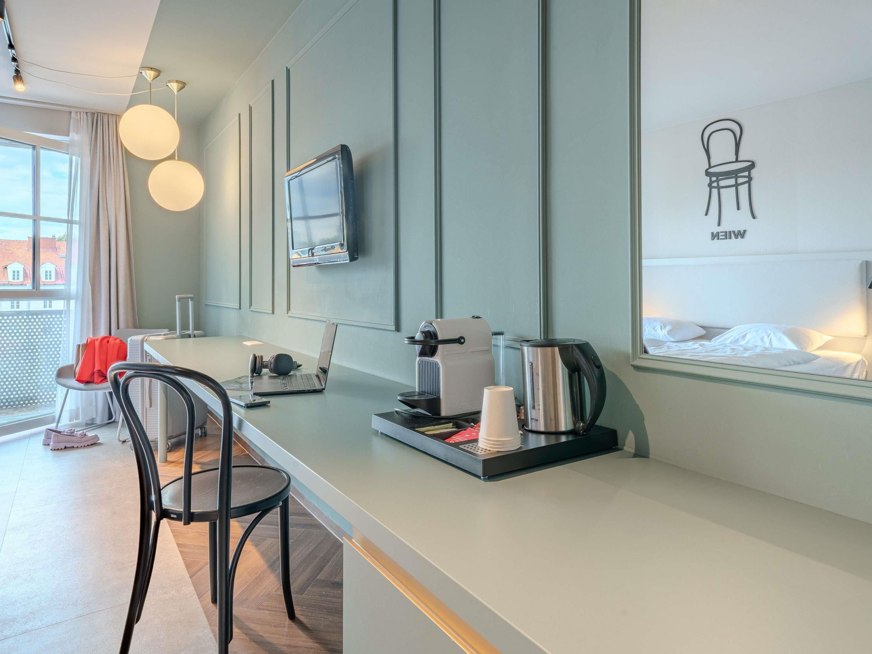 A modern hotel room with a kitchenette, a flat-screen TV mounted on the wall, a bed with white bedding, and a window with a view of buildings.