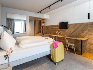 A hotel room with a bed, a desk, a chair, a TV, and a suitcase.