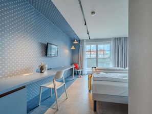 A hotel room with a bed, a desk with a chair, a television, and a window with curtains.