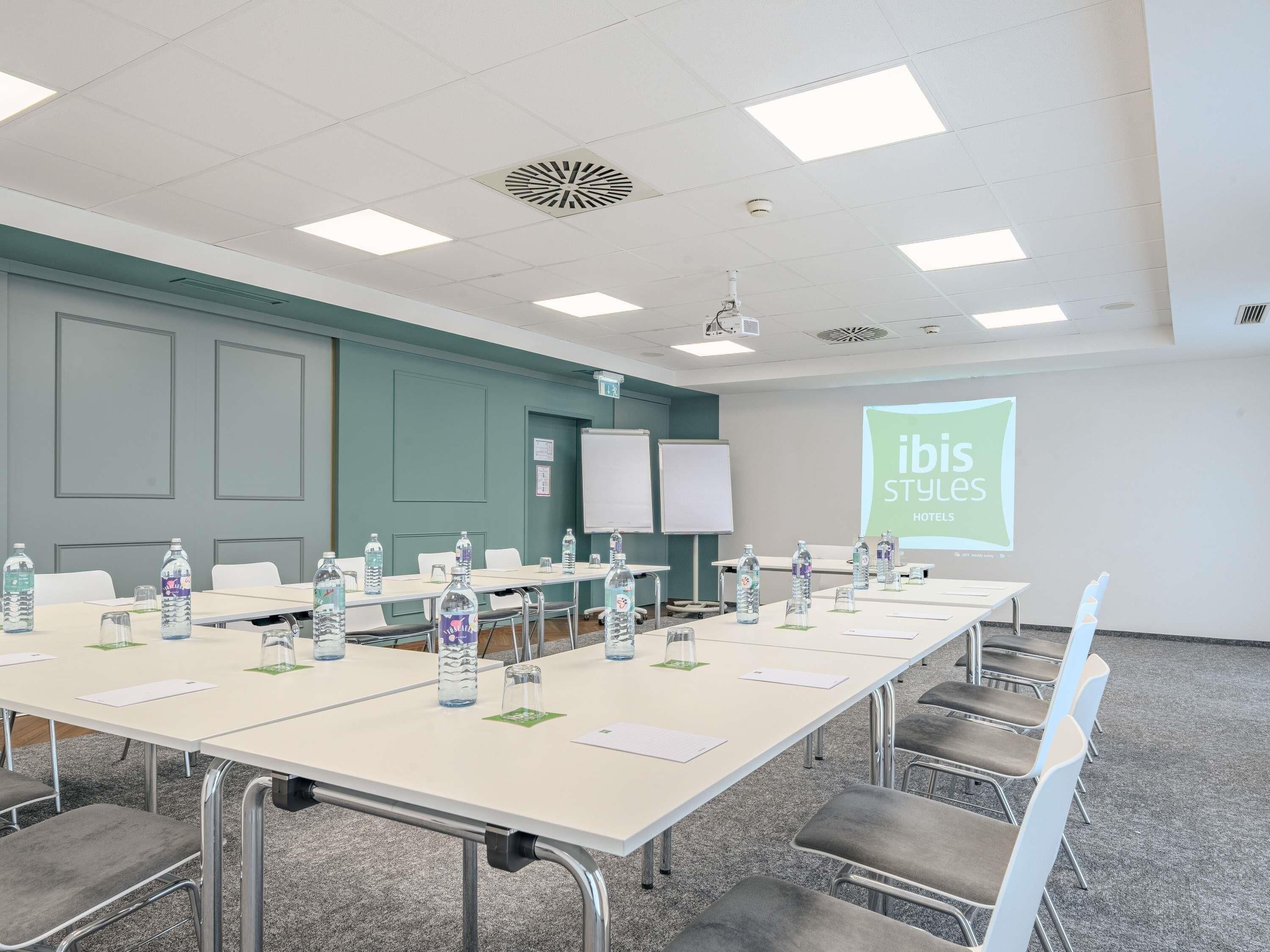 A conference room with a long table, chairs, water bottles, and a projector screen displaying 'ibis Styles Hotels'.