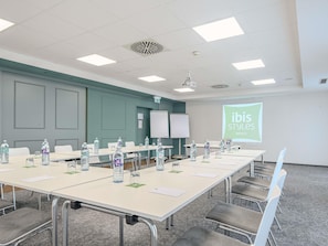 A conference room with a long table, chairs, water bottles, and a projector screen displaying 'ibis Styles Hotels'.