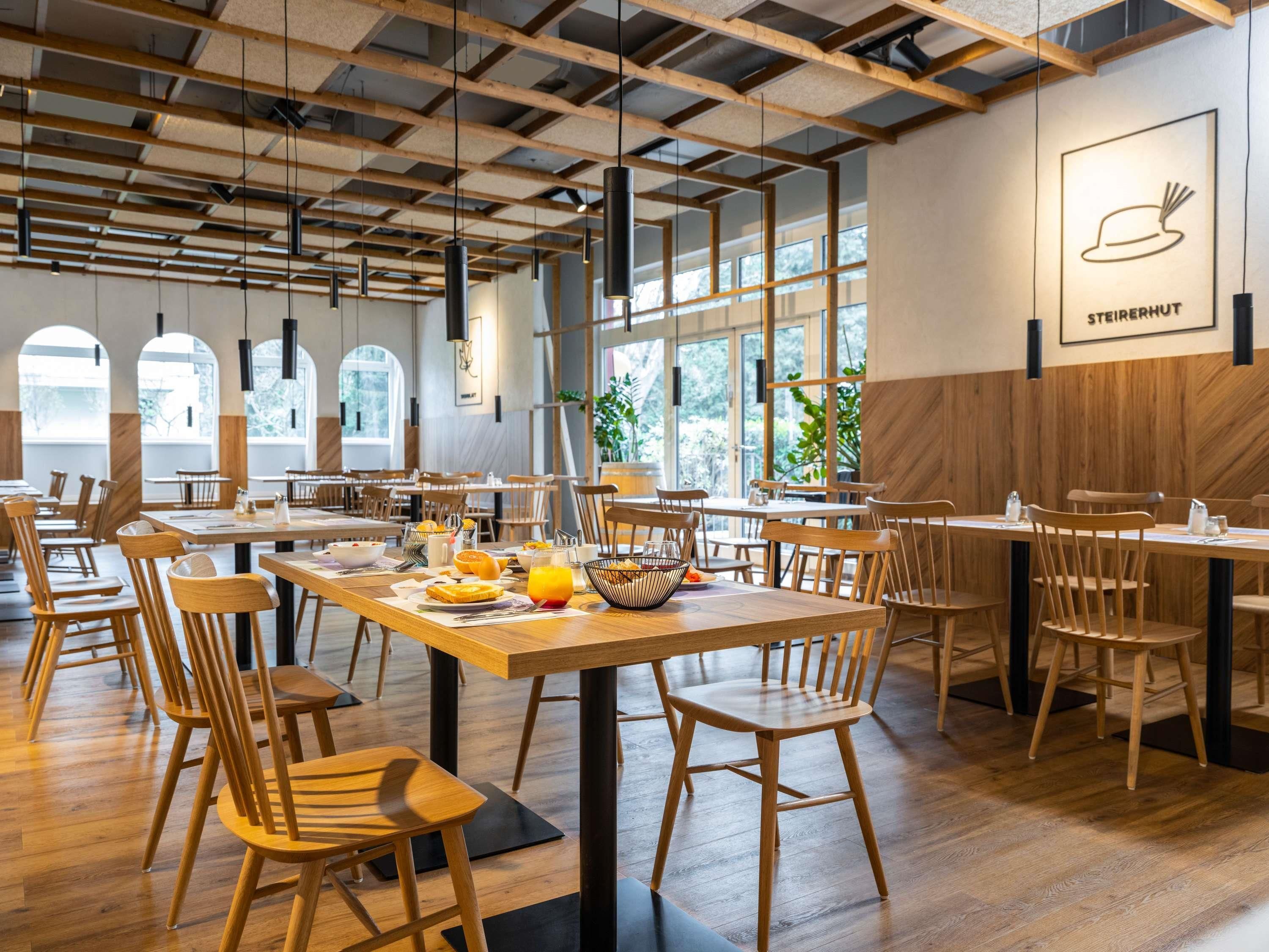 Un espace de restauration moderne avec des tables et des chaises en bois, de grandes fenêtres et une enseigne indiquant « STEIRERHUT ».