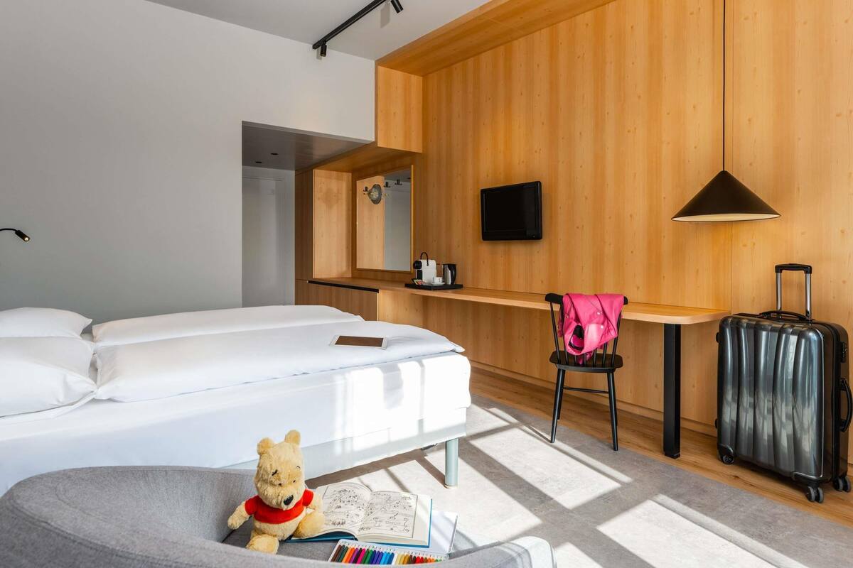 Quarto de hotel com cama, escrivaninha, cadeira, televisão, luminária, um urso de pelúcia, mala e janela.