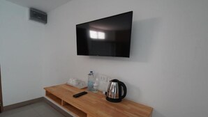 A room with a flat-screen TV mounted on the wall, a wooden desk with a kettle, a remote control, and a water bottle. - RoomQuest Tha Sala (Chiang Mai)