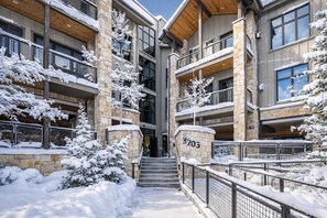 Un immeuble à plusieurs étages, avec une façade en pierre et en bois, des arbres couverts de neige et un escalier menant à l’entrée.