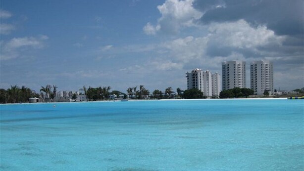 A coastal view with clear turquoise waters, a sandy beach, and tall residential buildings in the background.