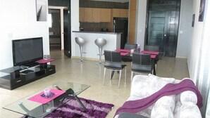 A modern living room with a glass coffee table, a flat-screen TV, and a sofa with a purple throw. There are two bar stools and a dining table with chairs. The kitchen is visible in the background with dark cabinets and a stainless steel refrigerator.