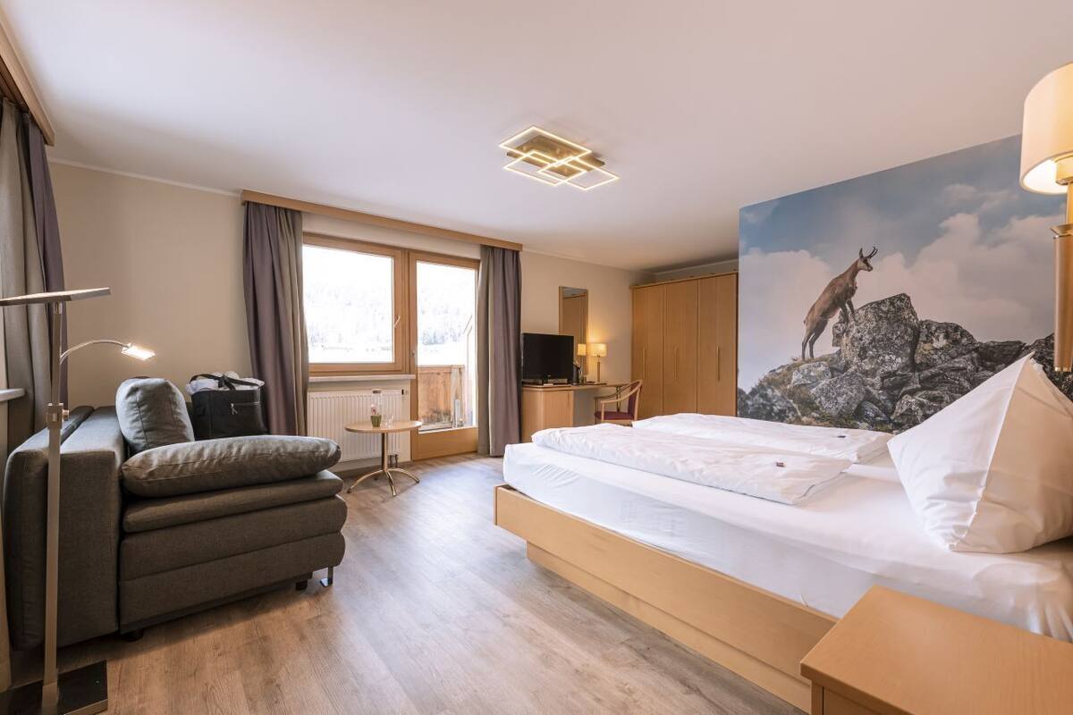 Et hotelværelse med seng, skrivebord, stol, sofa, fjernsyn og et maleri med et bjerglandskab.