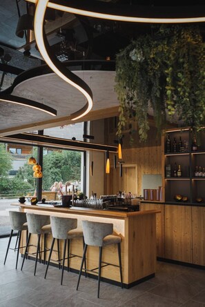Uma área de bar moderna com balcão de madeira, banquetas, plantas penduradas e vista para o exterior.