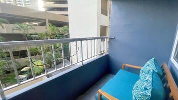 Un balcón con una pared azul, una silla de madera con cojín azul, y vistas a un edificio con varios pisos y balcones.