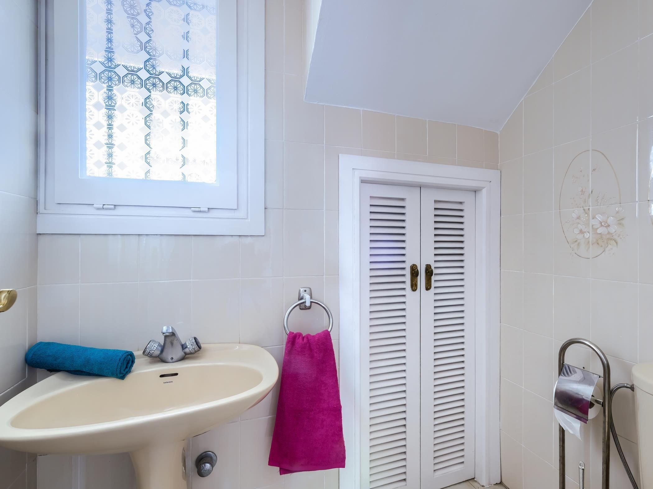 A bathroom with a sink, a window with a geometric pattern, a door with louvered panels, and a towel rack with a pink towel.