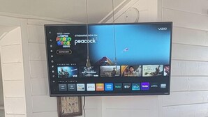 Een slaapkamer met een bed, een stoel, een aan de muur bevestigde televisie en een plafondventilator.
