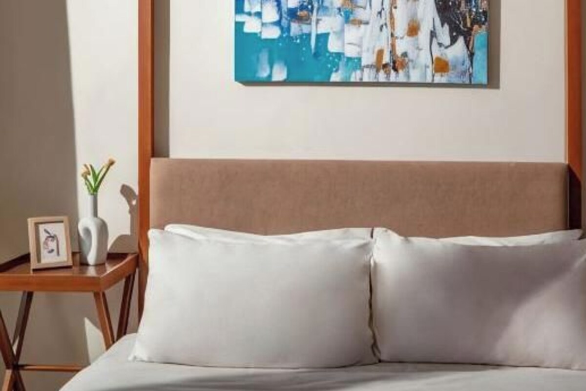 A bed with a headboard, two pillows, a bedside table with a vase and a picture frame, and a wall-mounted abstract painting.