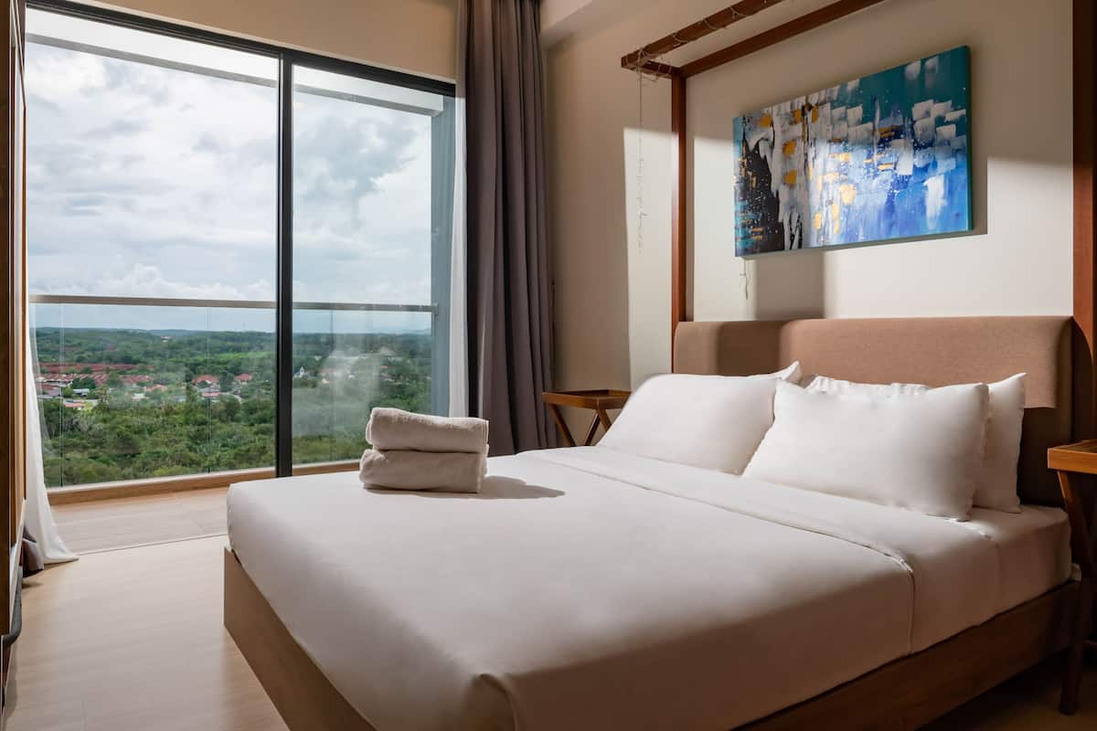 A hotel room with a large bed, a view of a cityscape through a floor-to-ceiling window, a painting on the wall, and a wooden nightstand.