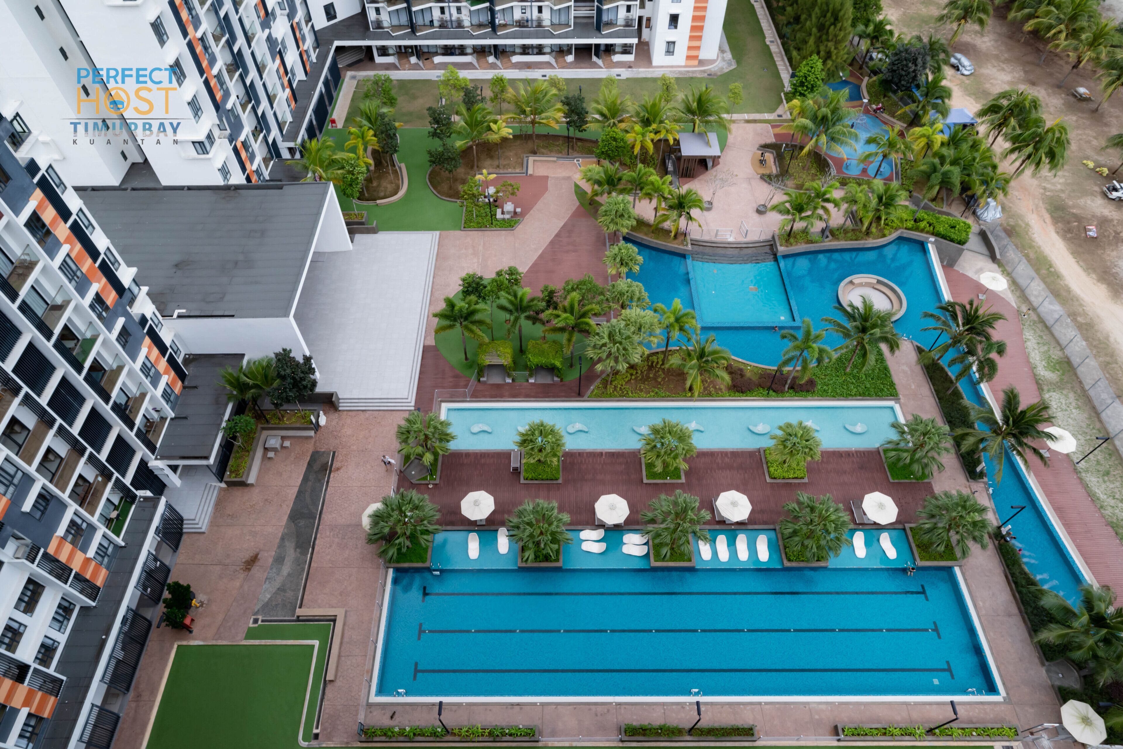 Et hotel med flere etager og et poolområde på taget, der inkluderer en stor swimmingpool og en mindre, rund pool, omgivet af palmer og grønt.