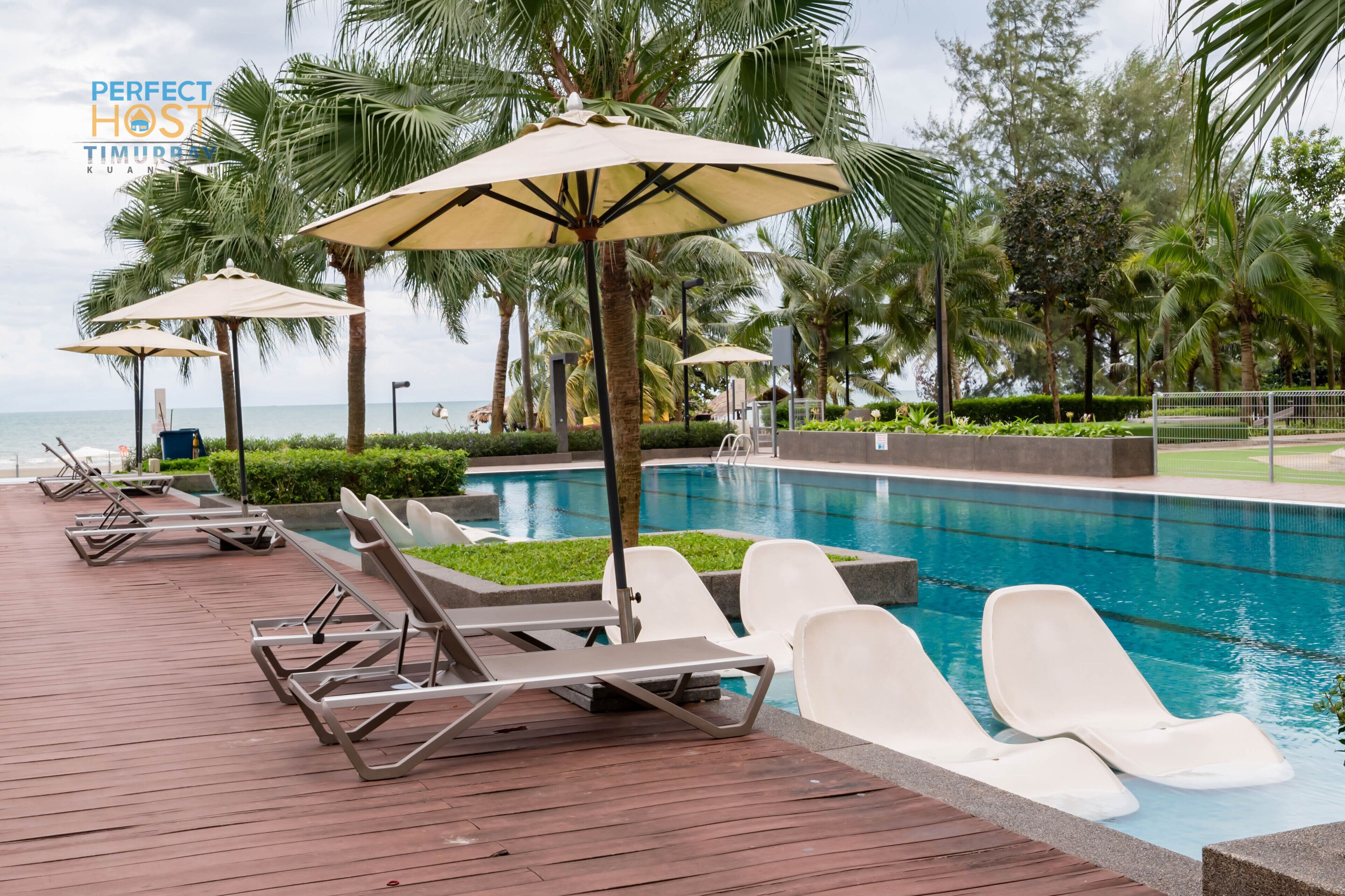 A poolside area with lounge chairs and umbrellas, surrounded by palm trees and a view of the sea.
