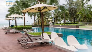 A poolside area with lounge chairs and umbrellas, surrounded by palm trees and a view of the sea.