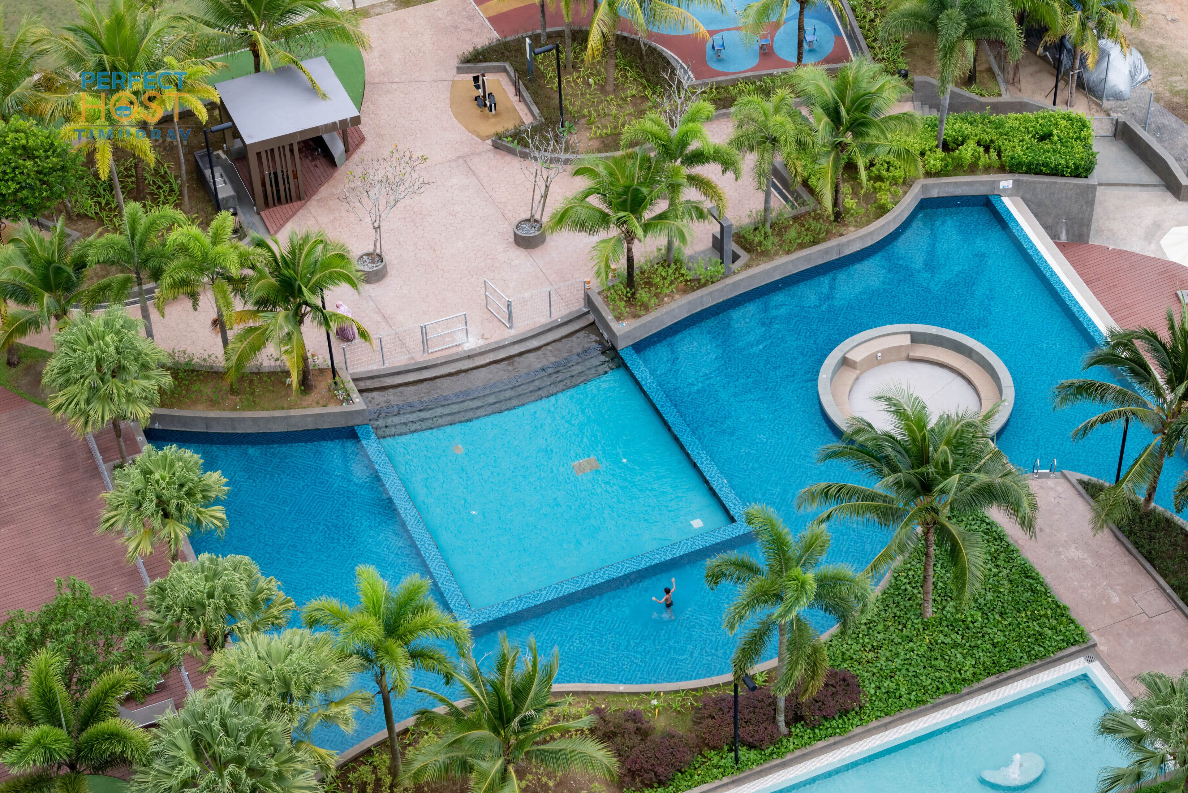 A large outdoor swimming pool surrounded by palm trees and greenery, with a small building and a basketball court nearby.