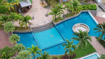 A large outdoor swimming pool surrounded by palm trees and greenery, with a small building and a basketball court nearby.