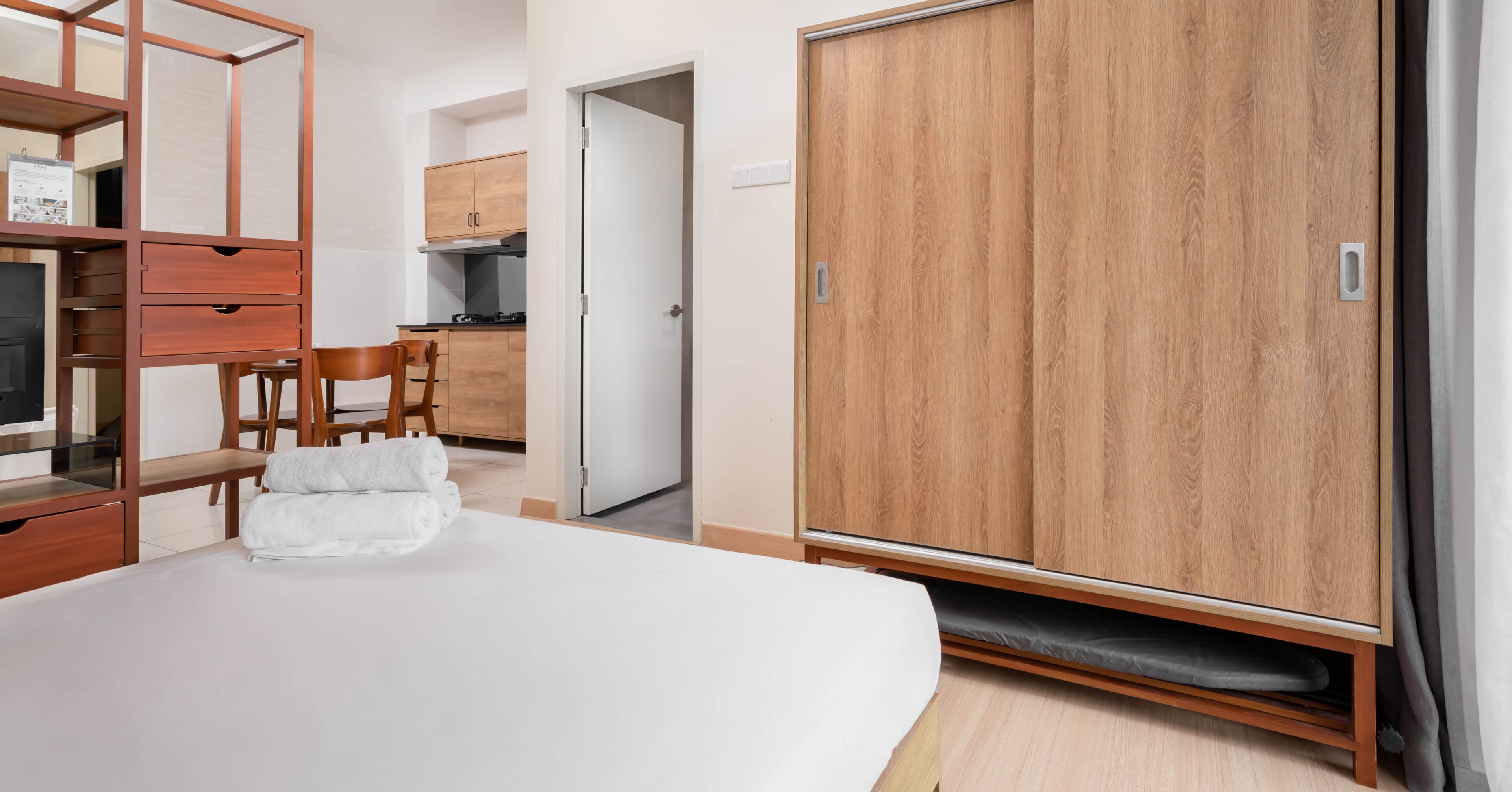 A bedroom with a bed, a wardrobe, a desk with drawers, a chair, and a small kitchen area with cabinets and a stove.