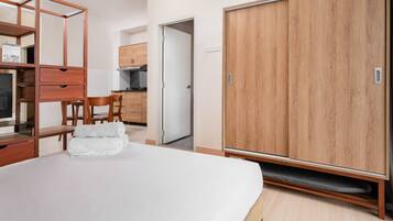 A bedroom with a bed, a wardrobe, a desk with drawers, a chair, and a small kitchen area with cabinets and a stove.