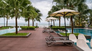 A poolside area with lounge chairs and umbrellas, palm trees, and a view of the sea. - Timurbay beachfront by Perfect Host (Kuantan)