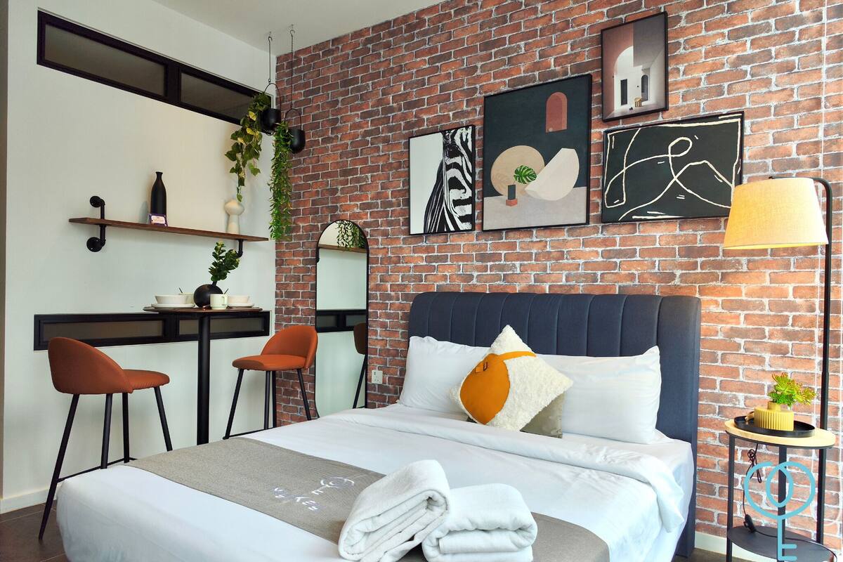 A modern hotel room with a brick wall, a bed with white linens, a desk with a chair, and artwork on the wall.
