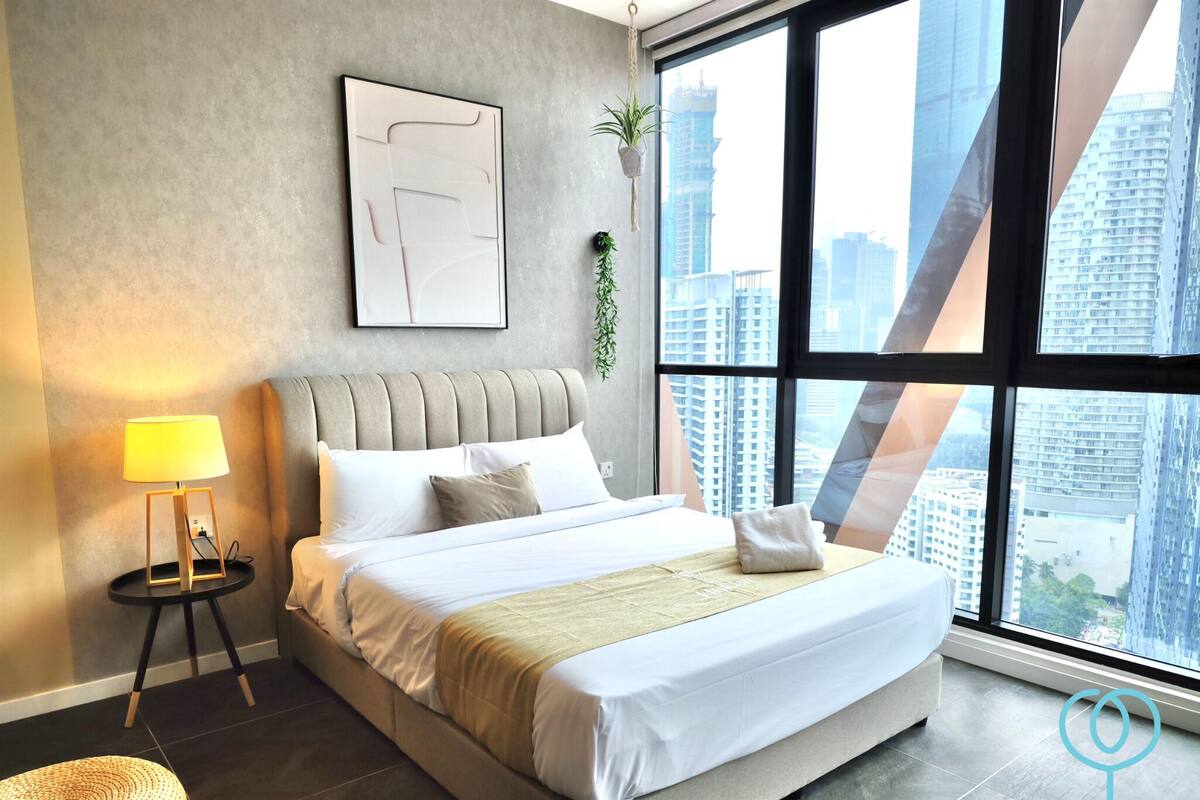 A modern hotel room with a large bed, a bedside lamp, a framed abstract artwork, and a view of the cityscape through large windows.