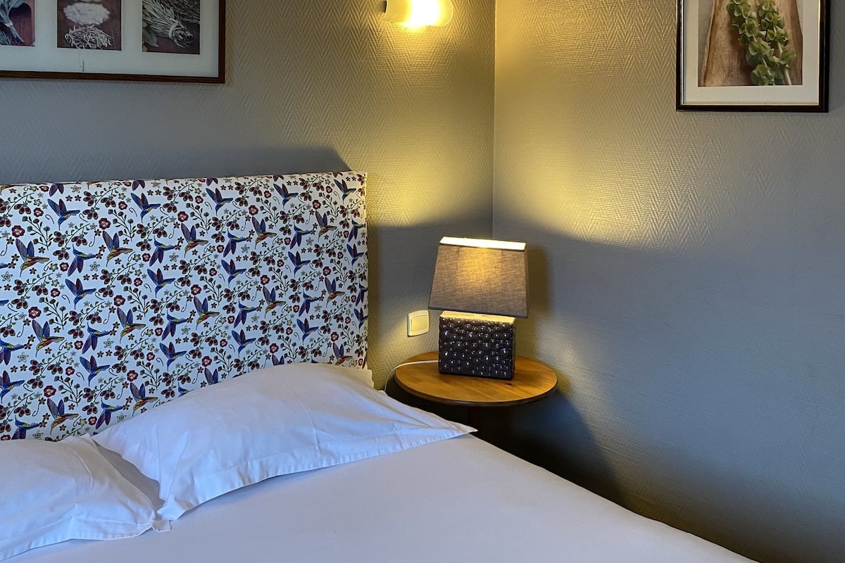 Una habitación de hotel con cama, una mesita de noche con lámpara, luz de pared y cuadros enmarcados.