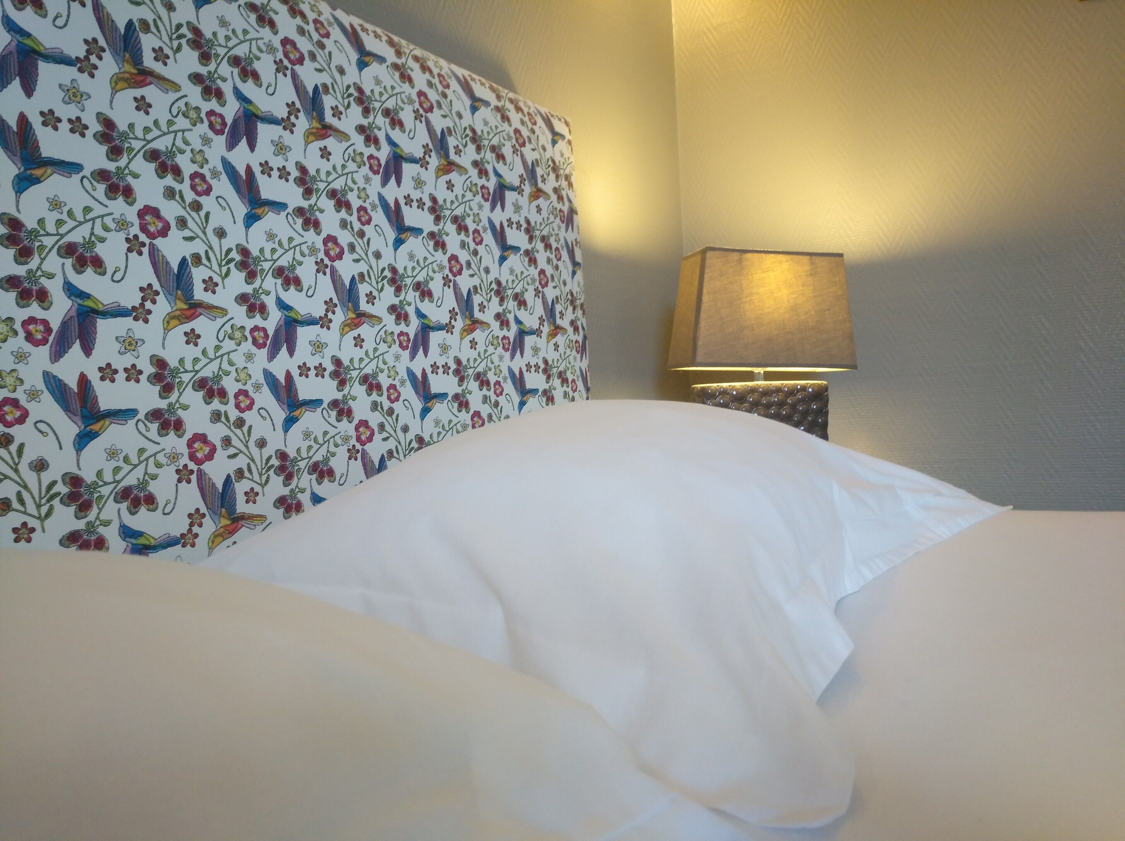 Un lit avec du linge de lit blanc et une tête de lit à motifs floraux. Une lampe se trouve sur une table de chevet.