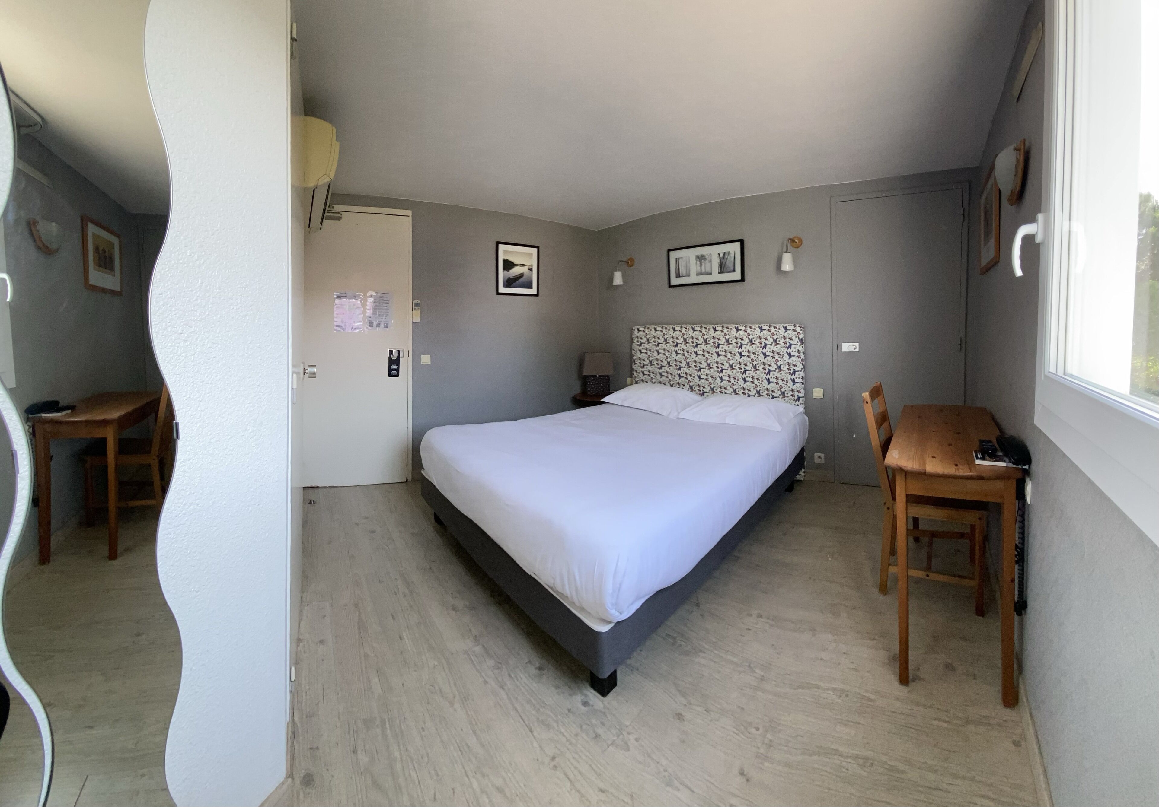 A bedroom with a bed, a desk, and a chair. There are framed pictures on the wall, a window, and a door.