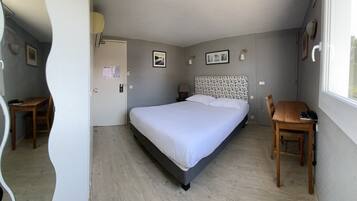 Una camera da letto con un letto, una scrivania e una sedia. Si vedono quadri appesi al muro, una finestra e una porta.