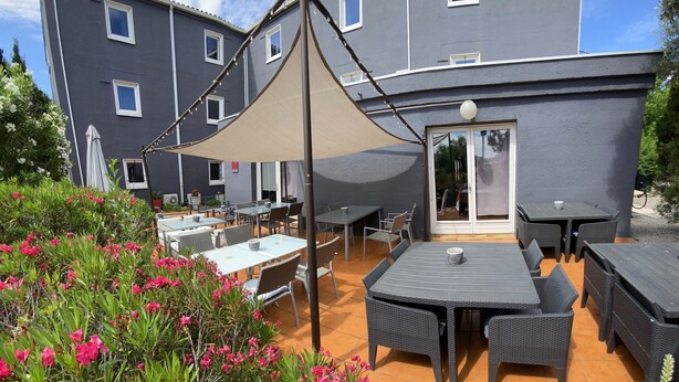 An outdoor patio area with multiple tables and chairs, surrounded by flowering plants and a building with large windows. - Le Sud Hotel (Mauguio)