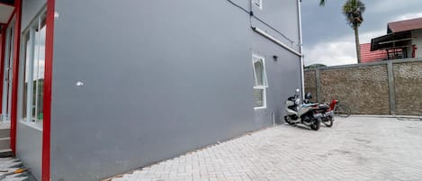 Um edifício com fachada cinzenta, entrada emoldurada por vermelho e um espaço pavimentado com uma motocicleta estacionada.