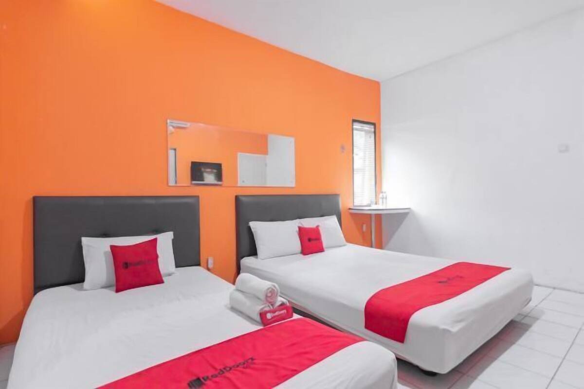 Une chambre d’hôtel avec deux lits, chacun agrémenté de draps blancs et de touches de couleur rouge, des murs orange, une petite table d’appoint et une fenêtre avec des stores.