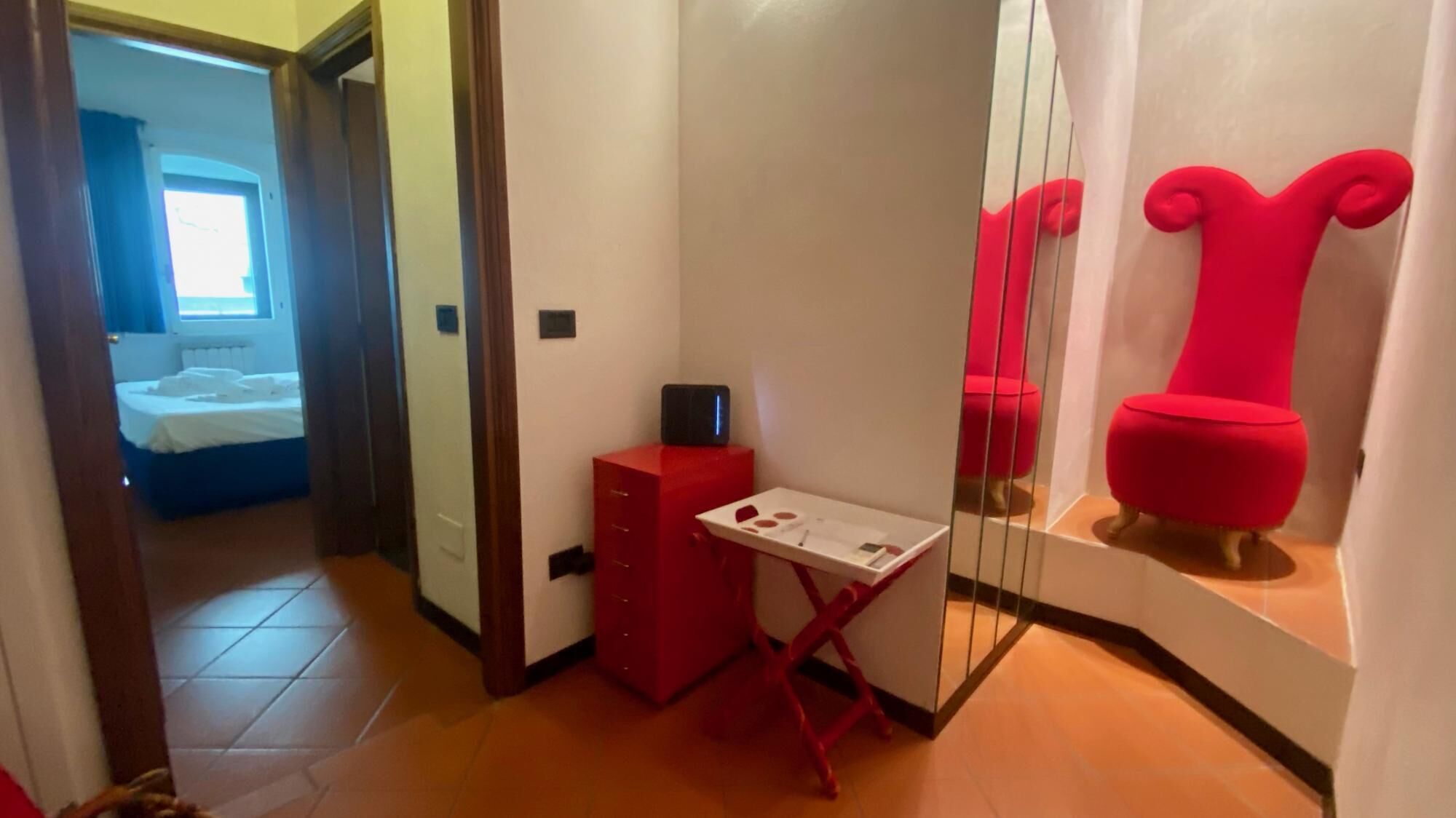 Habitación de hotel compacta con una cama, un pequeño escritorio con silla, un armario rojo y un espejo con detalle en rojo.