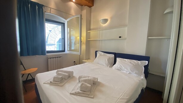 A hotel room with a bed, two pillows, a nightstand, a lamp, a window with curtains, and a towel on the bed. - Appartamento Lamberti (Florence)
