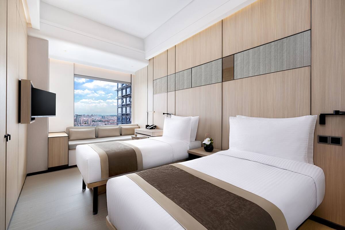 Une chambre d’hôtel moderne avec deux lits, une grande fenêtre donnant sur la ville, un téléviseur à écran plat et une petite table agrémentée d’un vase de fleurs.
